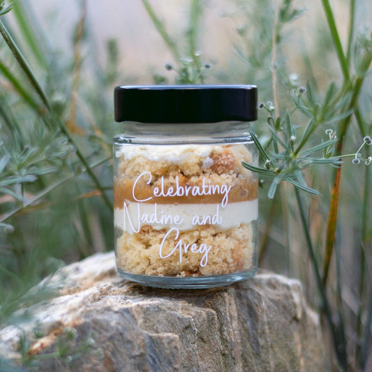 layers of cake and frosting in a jar for a birthday party with custom label for wedding guests saying celebrating nadine and greg